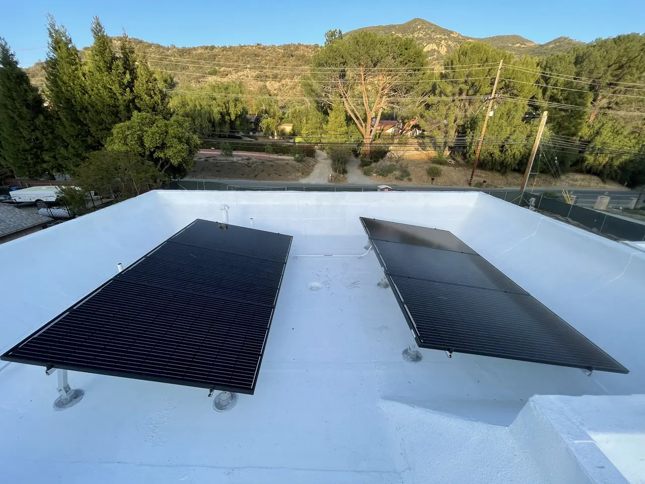 Two groups of black solar modules on low legs on a white flat roof, with trees and hills beyond, Calabasas