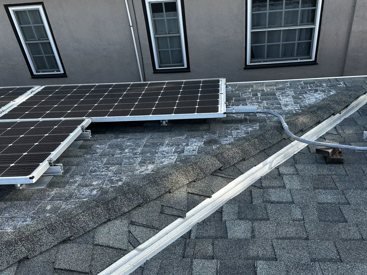 New solar modules mounted along a stucco wall with rail, conduit and a junction box on the shingle roof