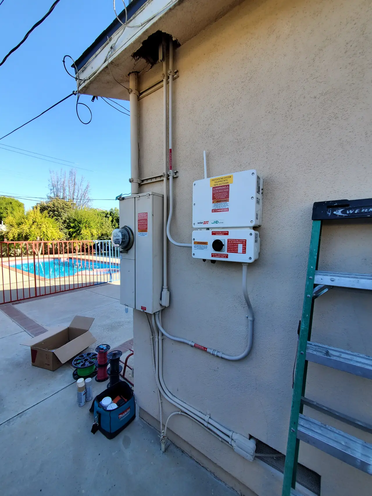 SolarEdge string inverter, safety switch and conduit on a stucco side wall beside a pool fence, Encino
