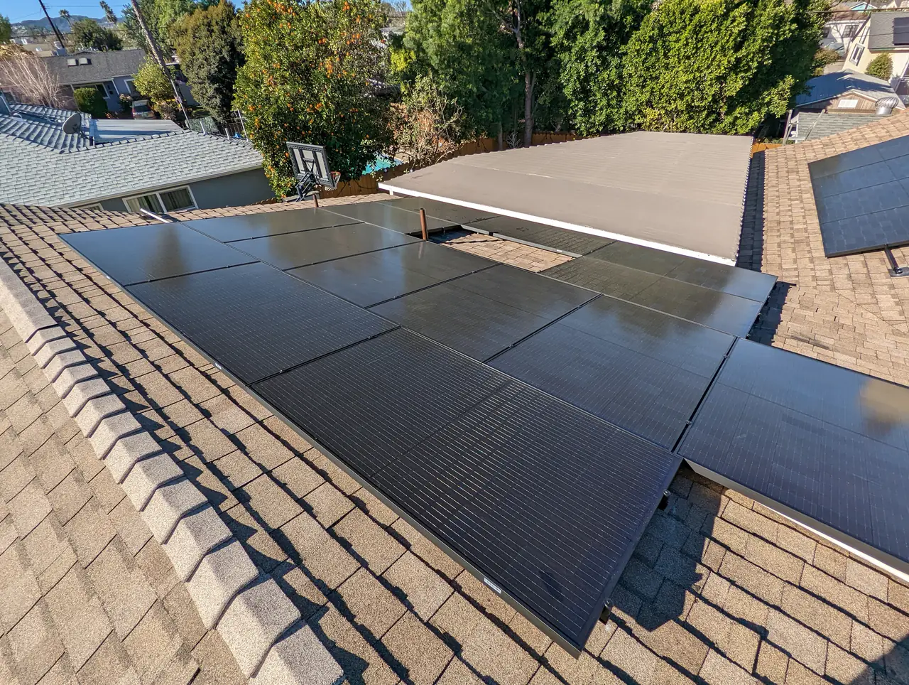 Black solar modules across several planes of a composition shingle hip roof in Encino, with trees and neighboring roofs beyond