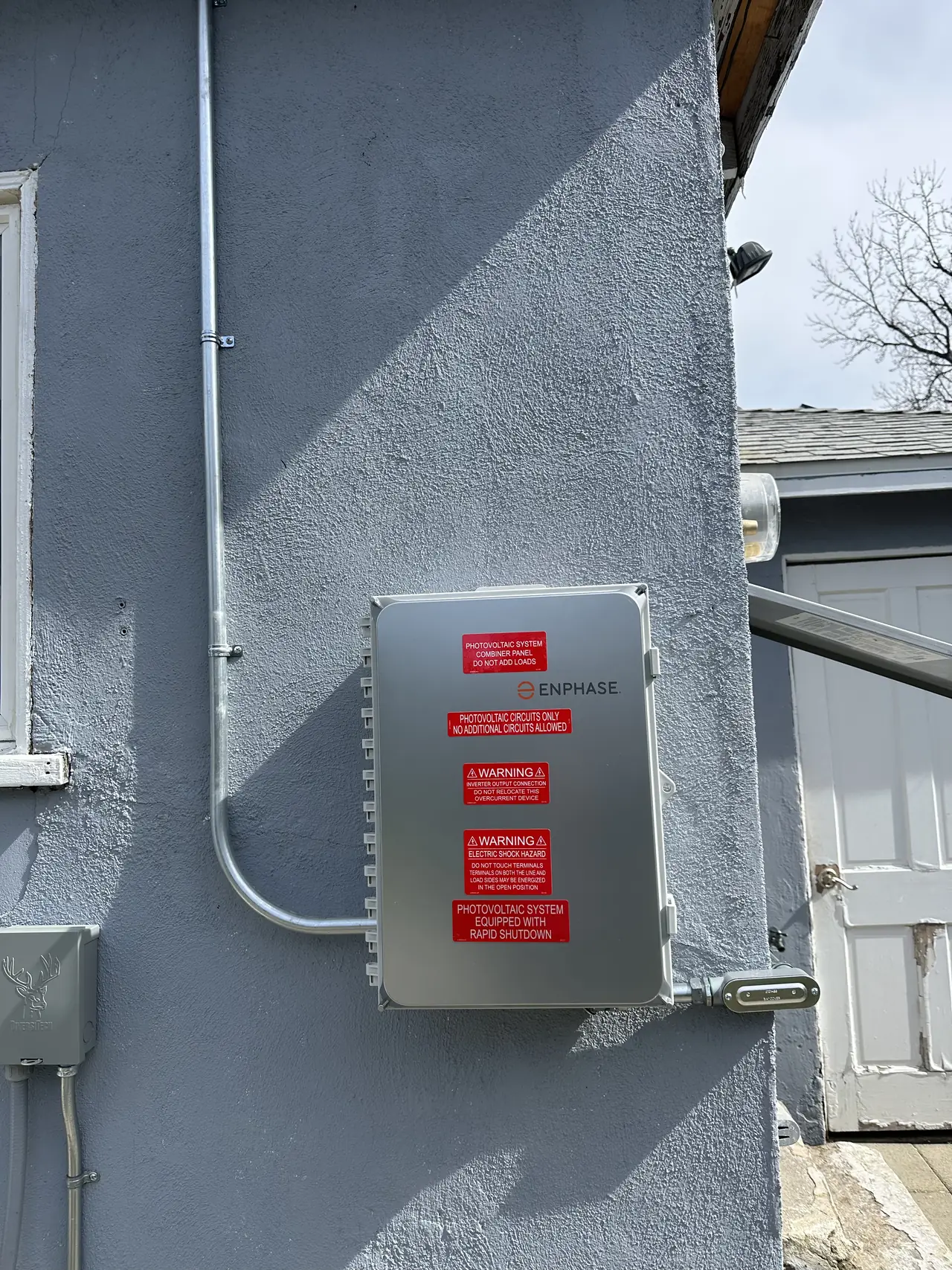 Enphase combiner panel with warning labels on a stucco wall, with conduit running to the roof