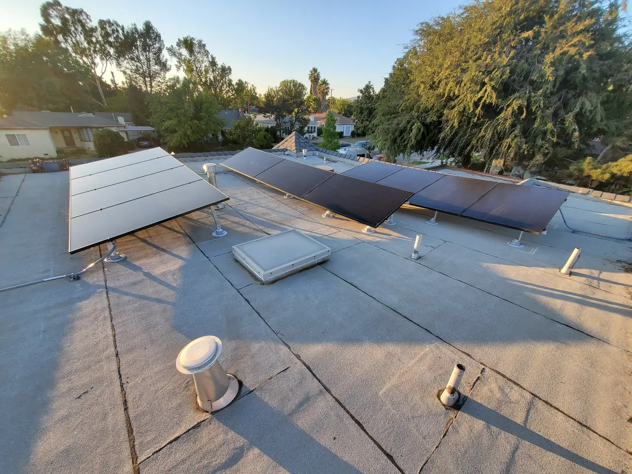 Three groups of tilted solar modules on a flat roof at sunrise with trees and rooftops beyond, Glendale