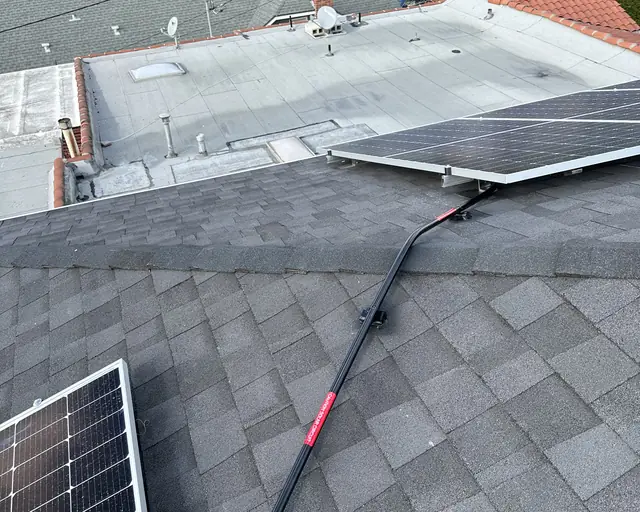 Black conduit running across the ridge of a gray composition shingle hip roof between two groups of solar modules, with a flat-roofed neighbor beyond, Glendale