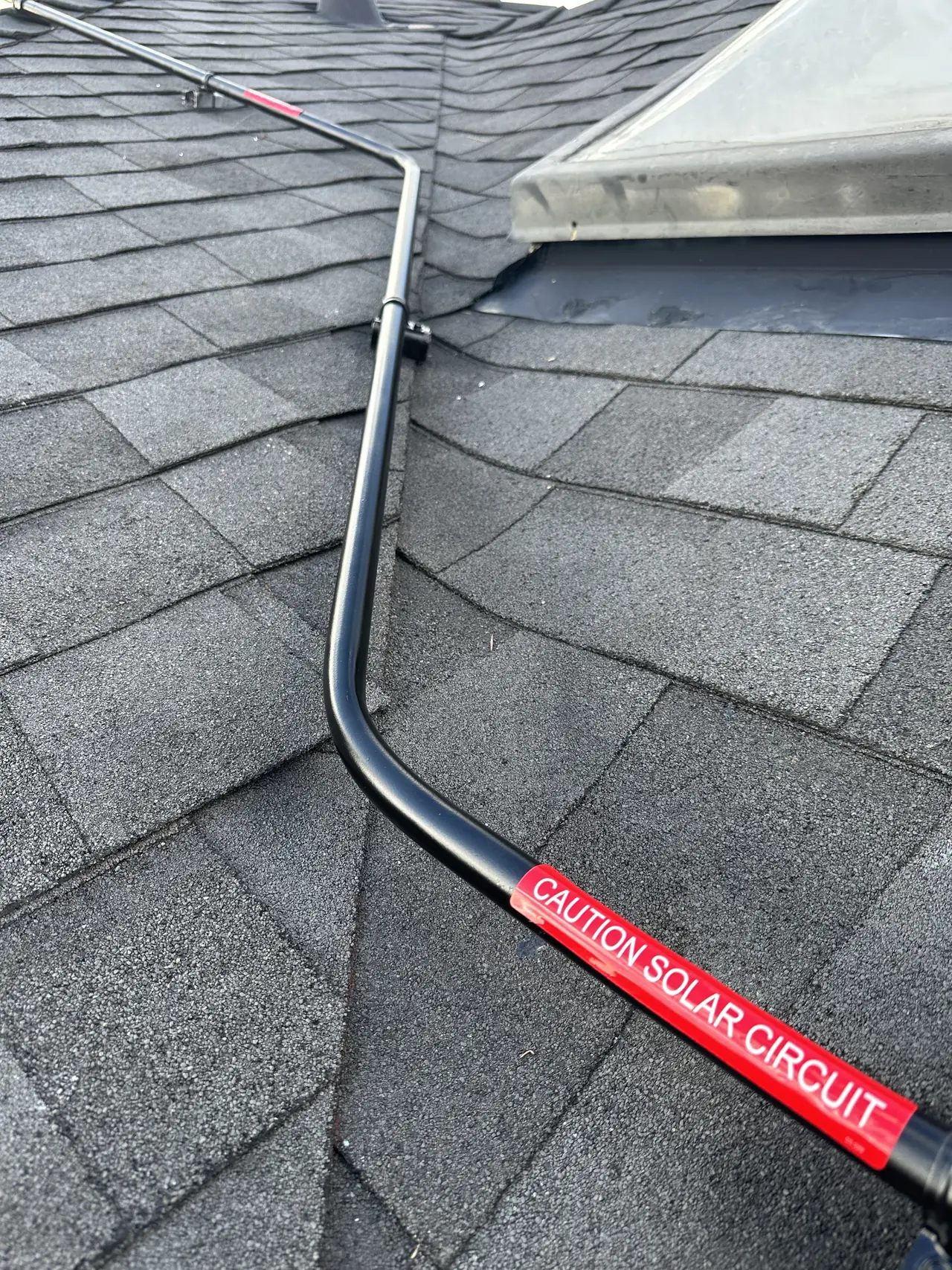 Black conduit with a red solar-circuit warning label crossing a shingle ridge beside a skylight, Glendale