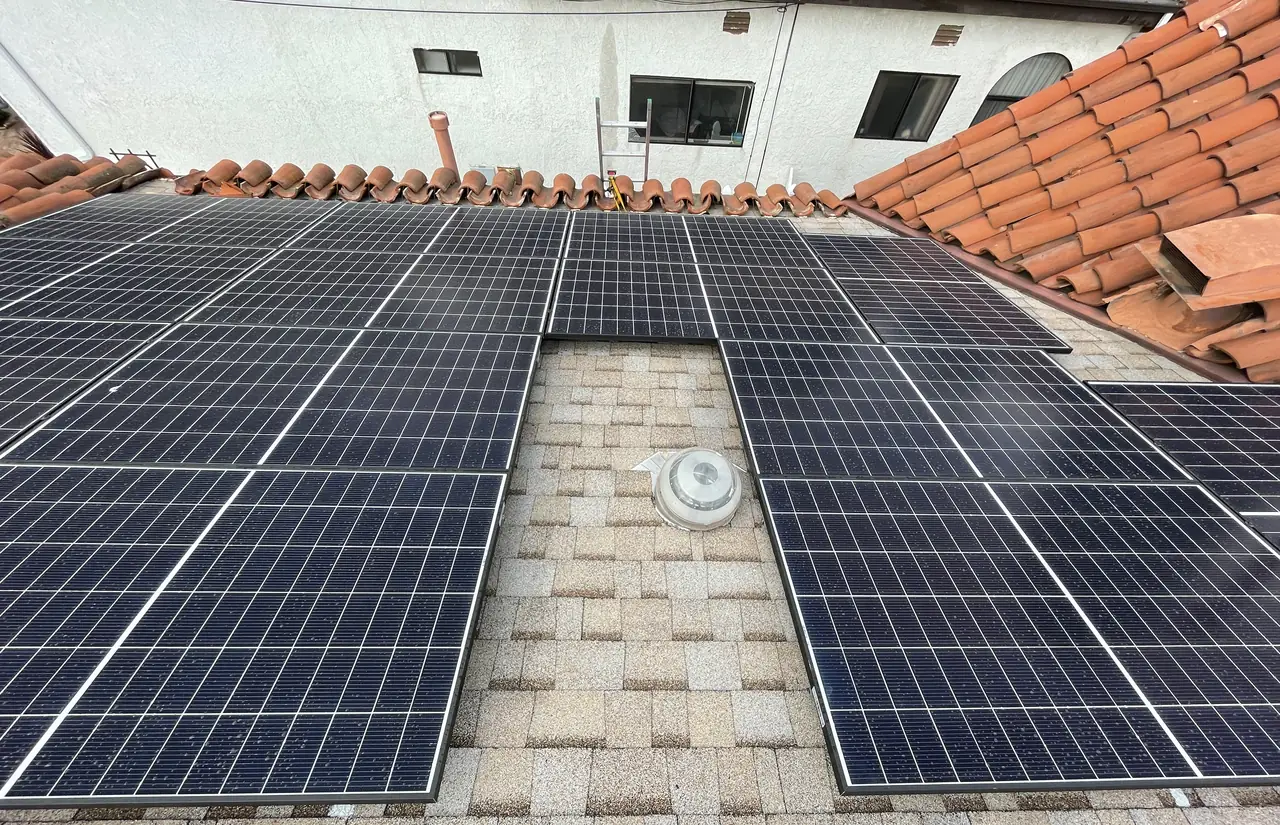 Two groups of solar modules on a shingle roof section with a gap left around a vent, bordered by clay tile, with a stucco wall beyond, Glendale