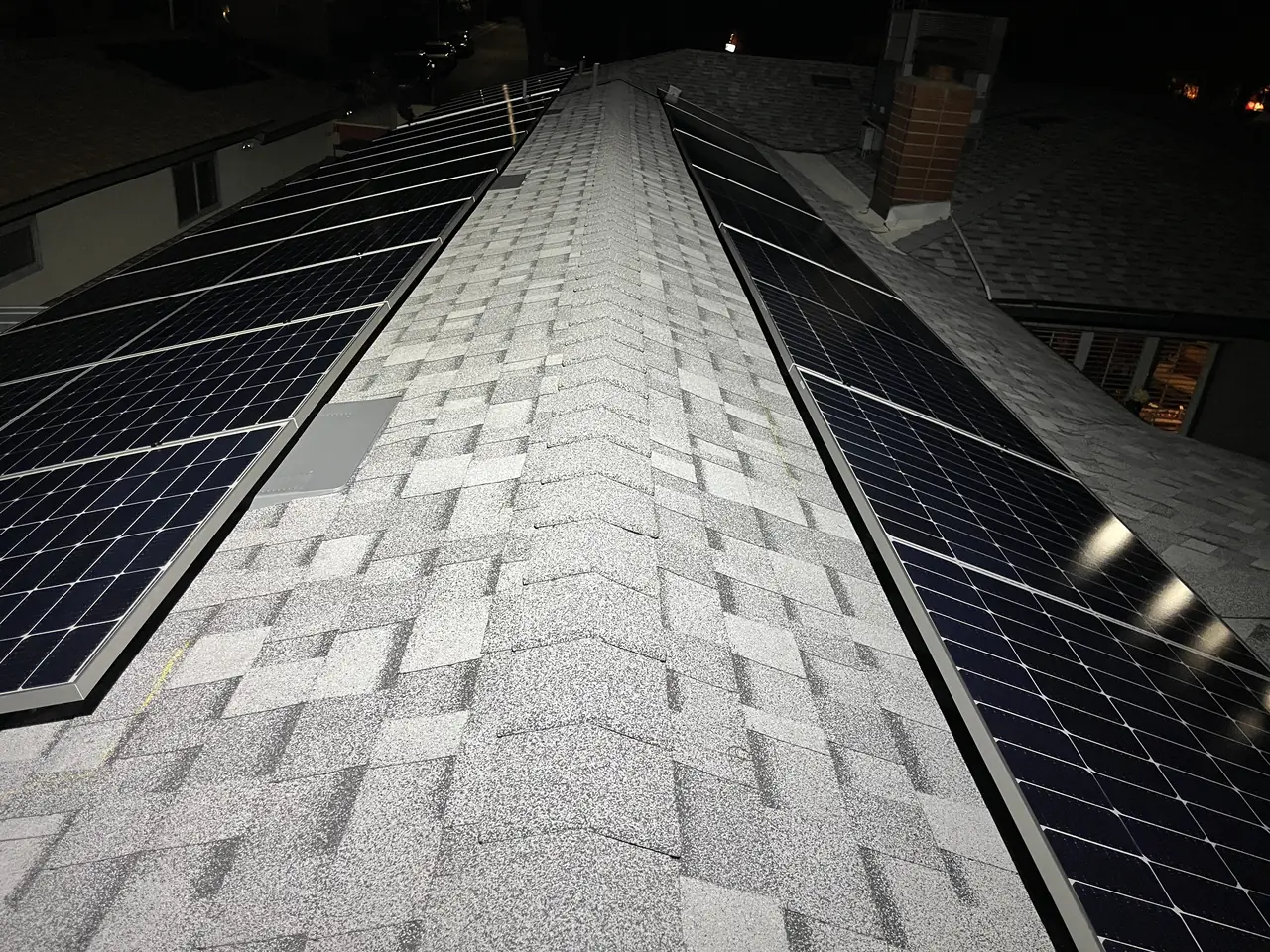 Two rows of solar modules flanking the hip of a composition shingle roof, with a chimney and neighboring rooftops beyond, Glendale