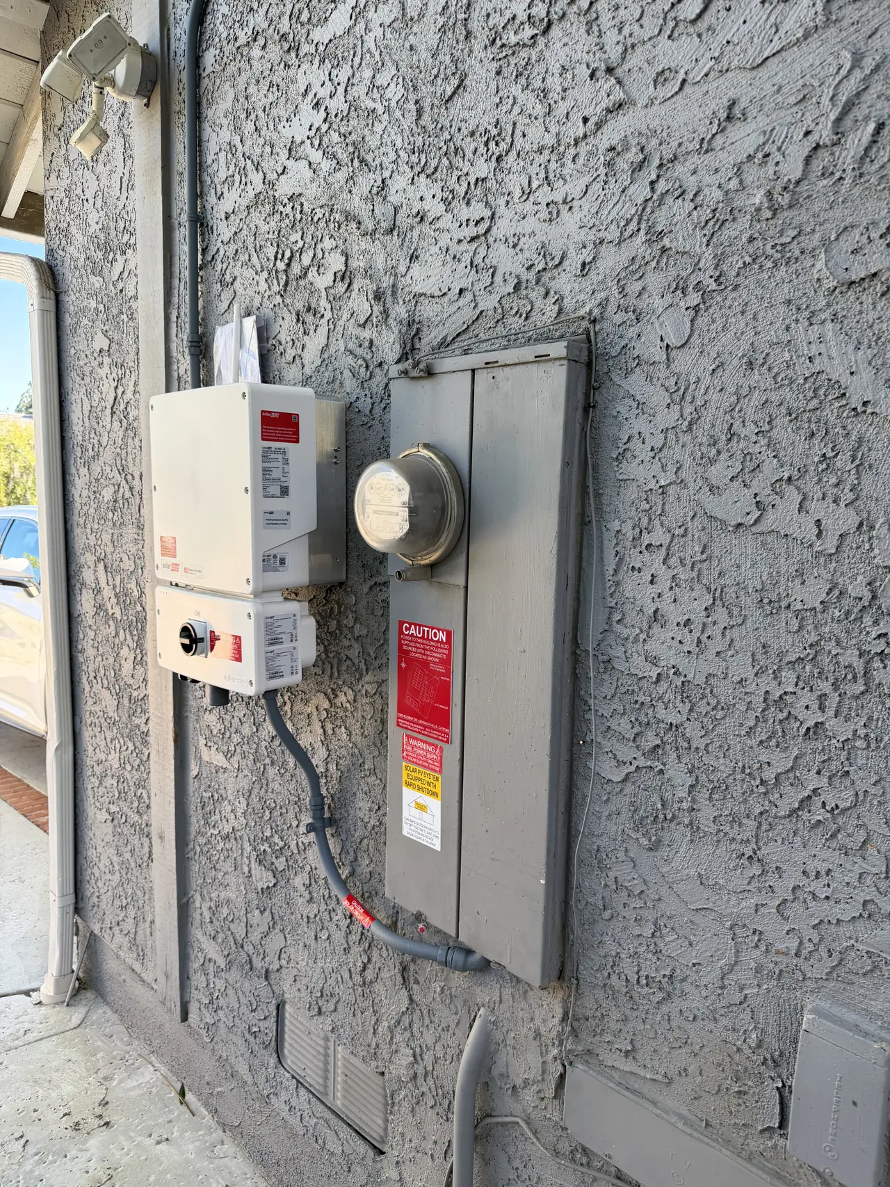 A string inverter, its disconnect and the closed service panel on a stucco wall with warning placards, Granada Hills