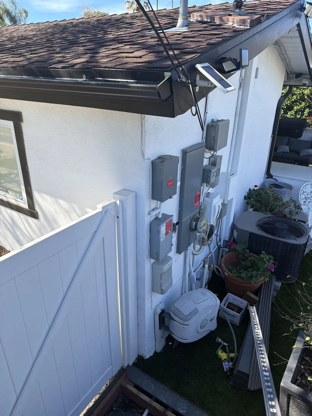Electrical equipment mounted on the white exterior wall of a dwelling beside a fence and planters, Granada Hills