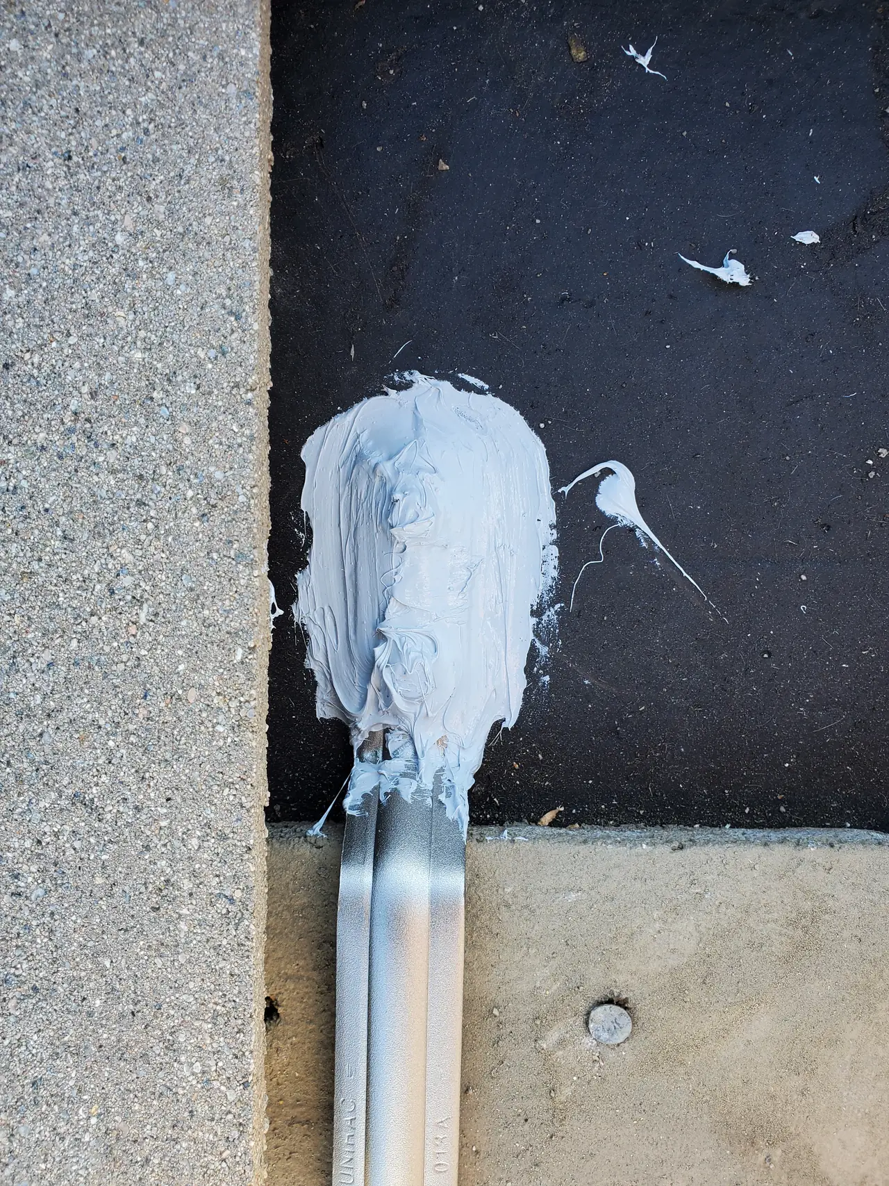 Gray mastic packed around the end of a mounting rail where it crosses the black membrane strip between concrete tiles, Granada Hills