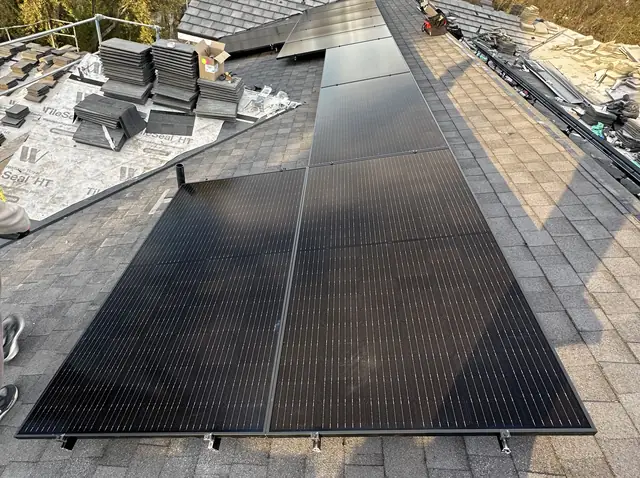 Black solar modules in a long row on a dark shingle roof of a new house, with flat concrete tile stacked on the next roof, Hidden Hills