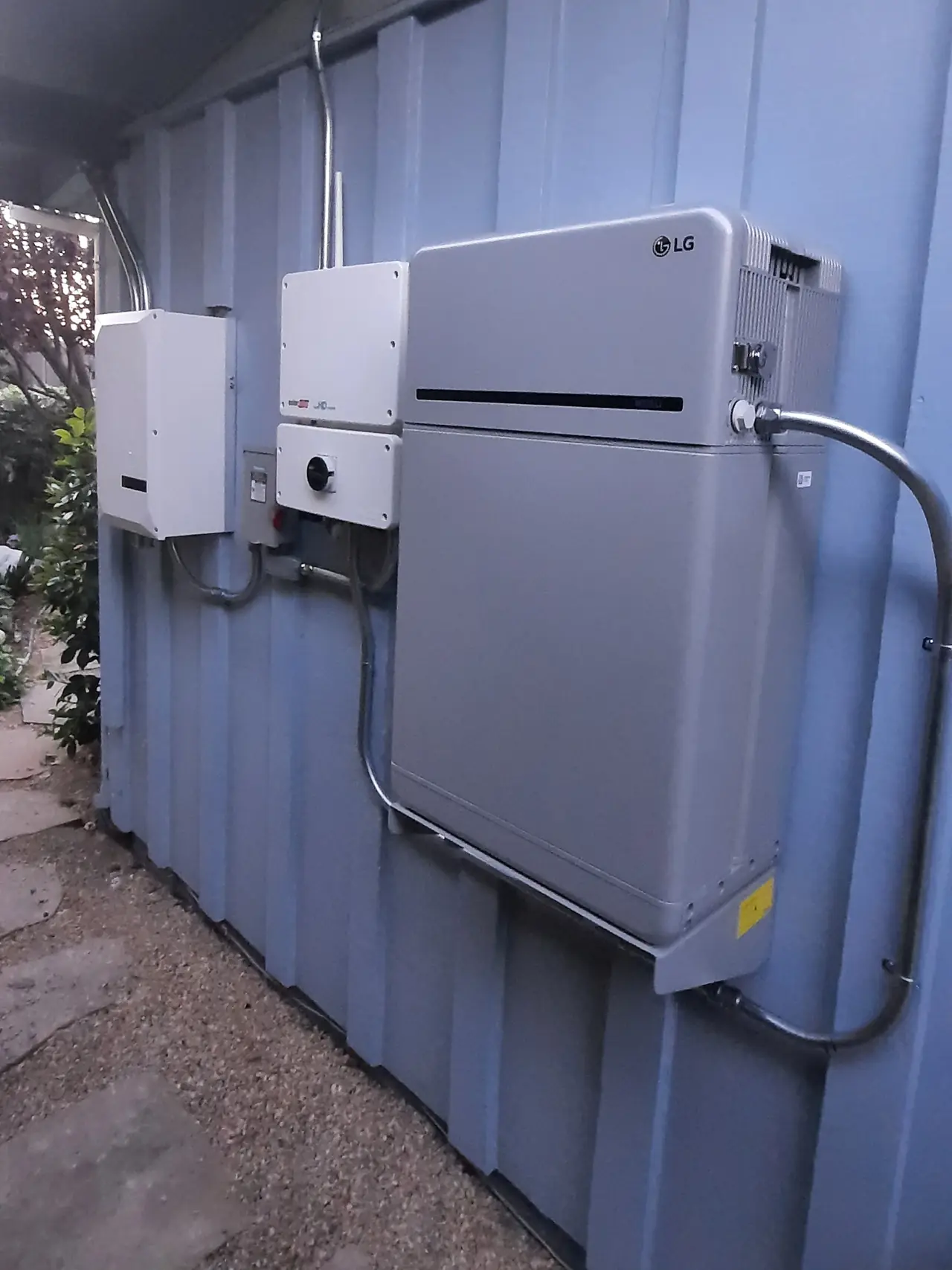 A wall-mounted battery beside an inverter and a disconnect under the eaves of a low house, Long Beach