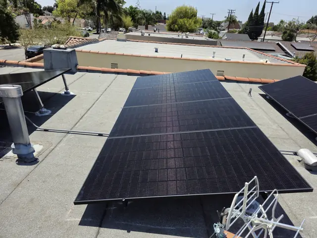 Tilted solar modules raised above a flat roof with a tile parapet and the neighborhood behind, Long Beach