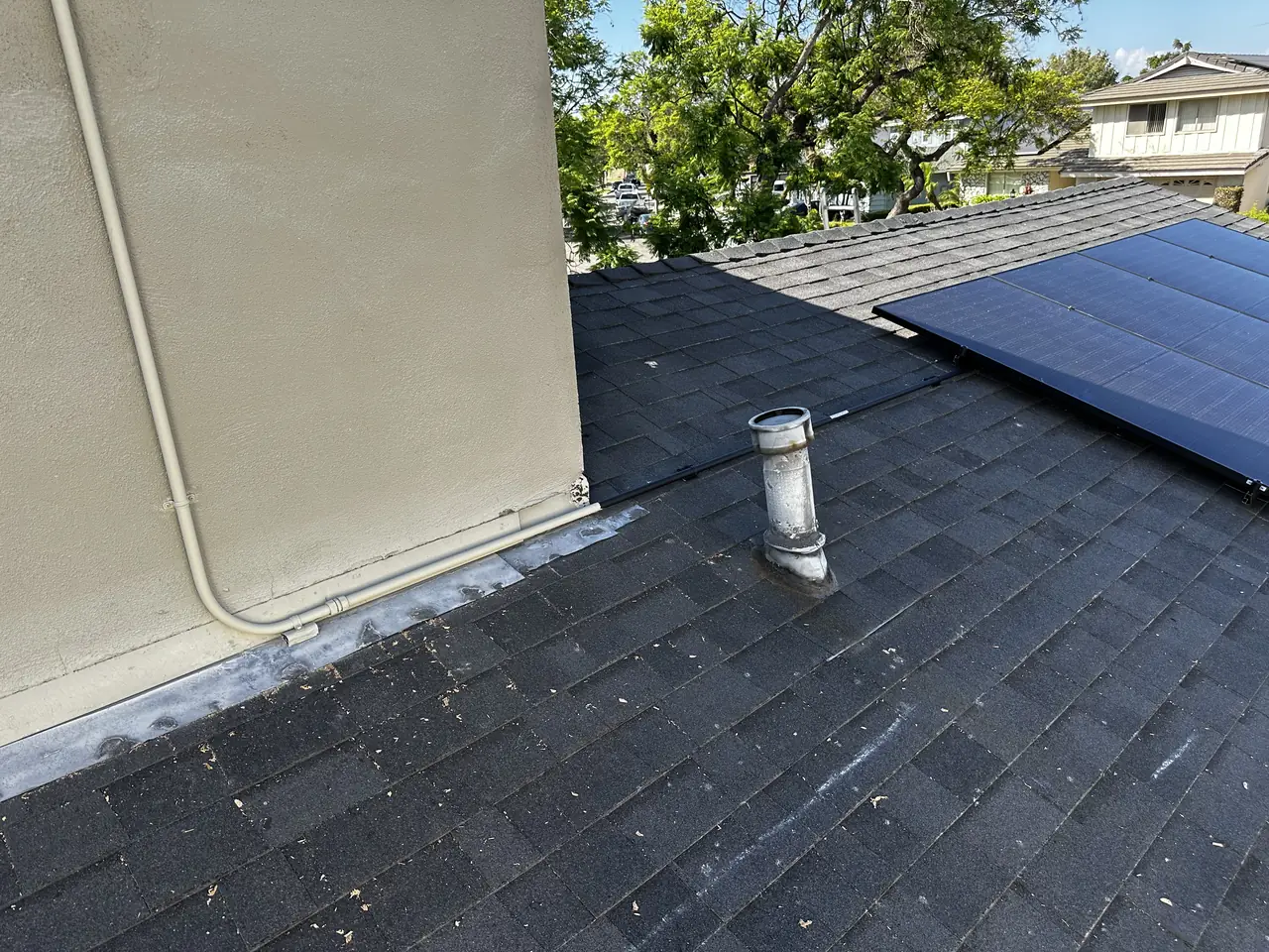The side edge of a solar array on a shingle roof beside a stucco wall and a roof vent, Long Beach