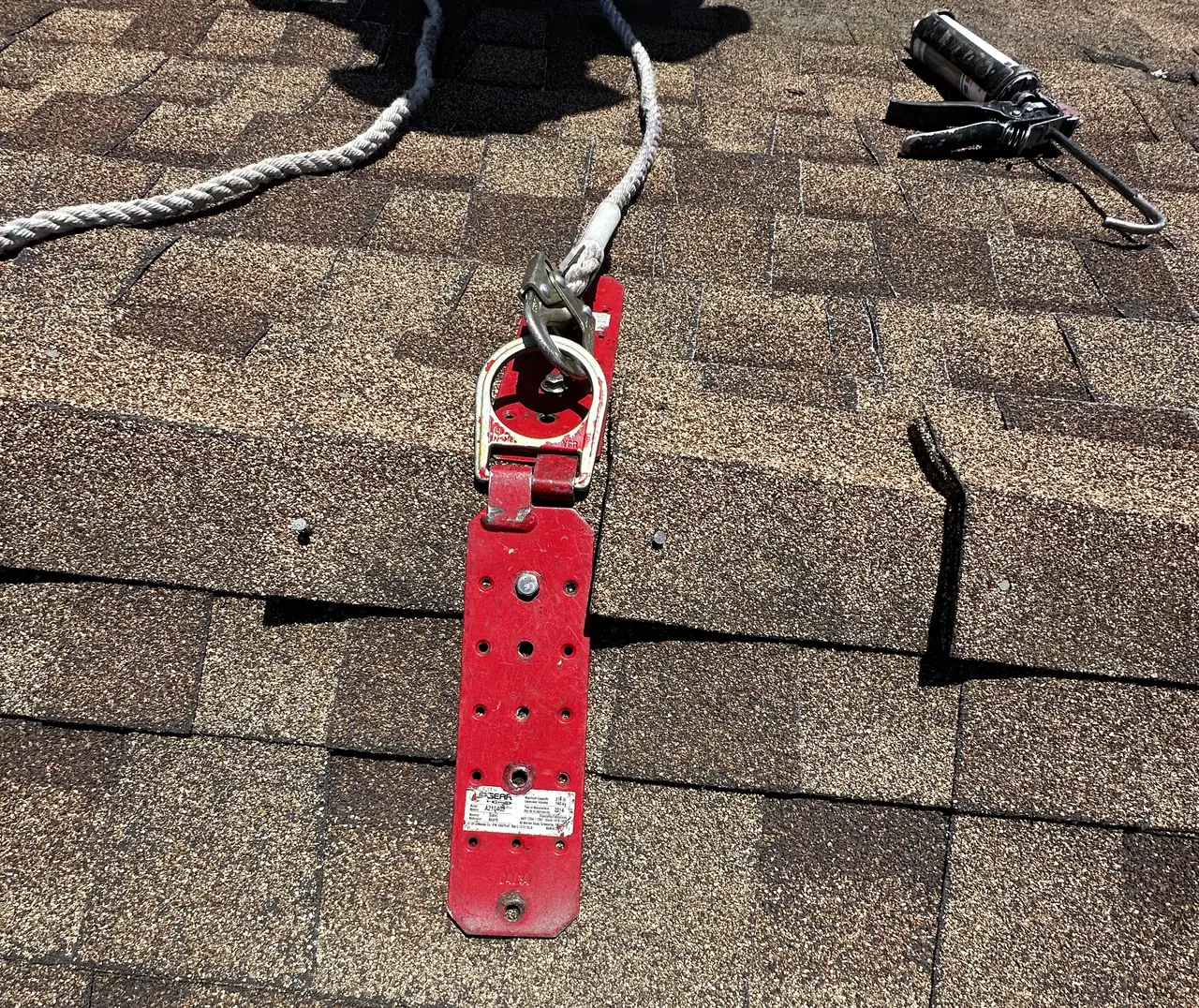 A red fall-protection anchor plate lagged through composition shingle with a lifeline clipped to its ring, Long Beach