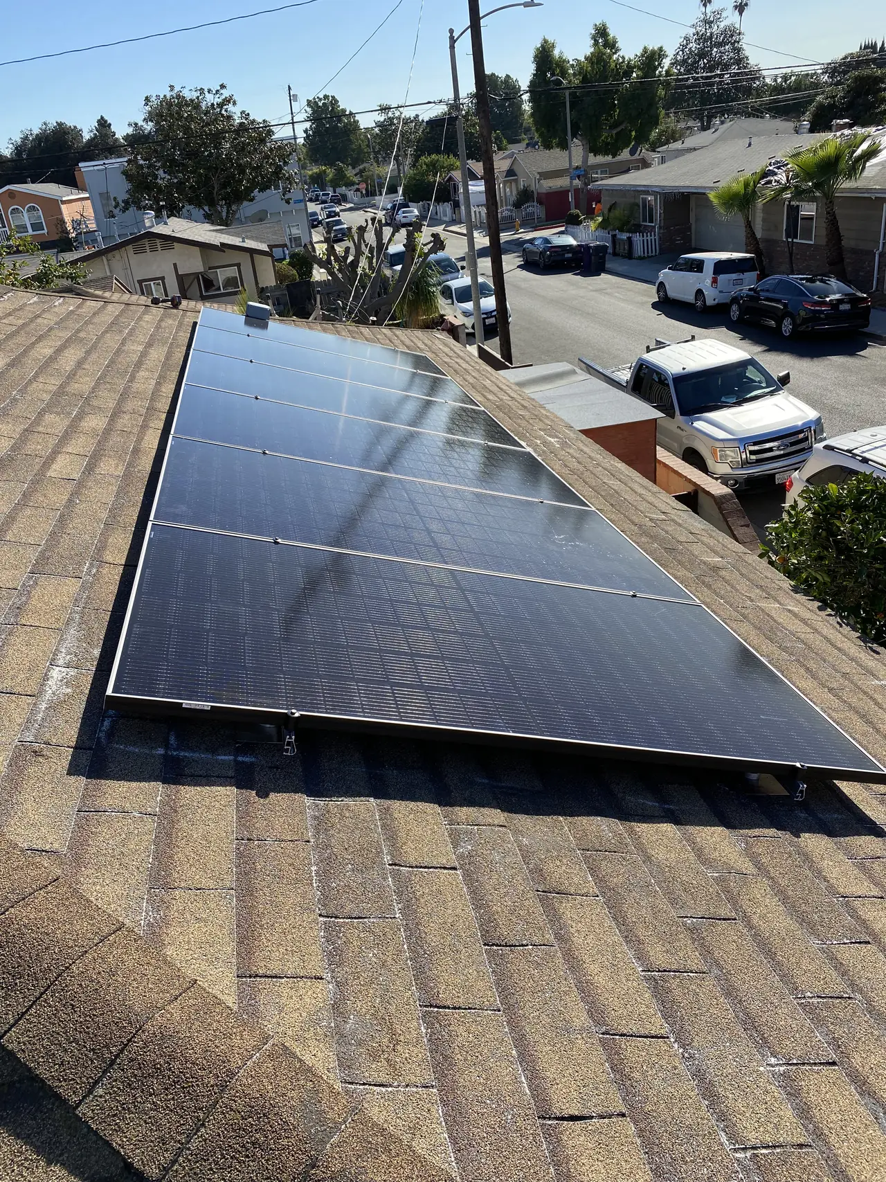 A short run of solar modules on a shallow shingle roof with parked cars and the street beyond, Long Beach
