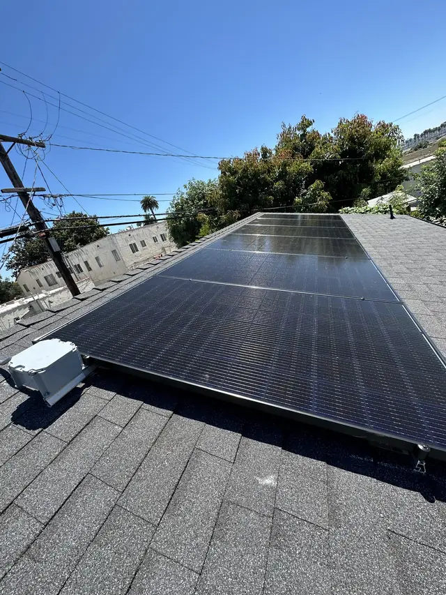 A small solar array on a composition shingle roof with trees and blue sky, West Adams, Los Angeles