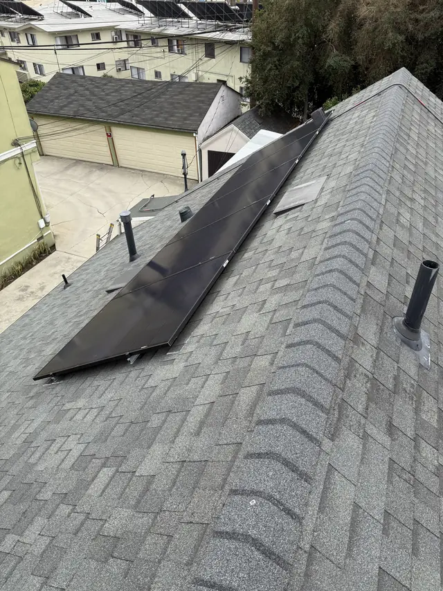 Black solar modules on a composition shingle slope above a pitched roof ridge, with neighboring rooftops beyond, Mid-City, Los Angeles