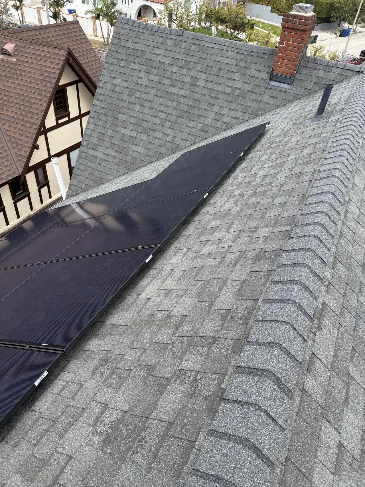 A solar array on a composition shingle roof seen along the ridge, with a brick chimney and tiled neighboring roof behind, Mid-City, Los Angeles