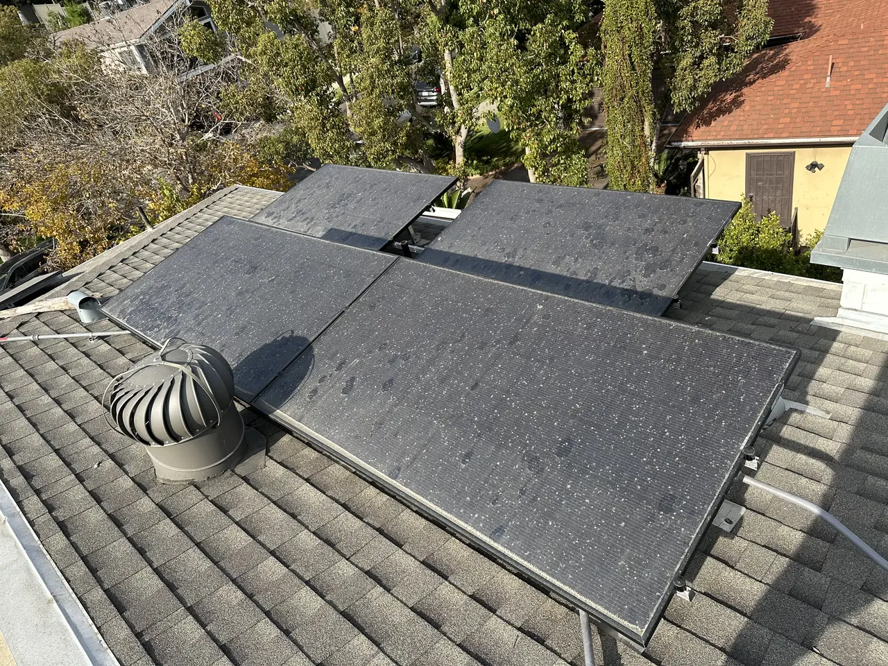 A solar array on a composition shingle roof beside a turbine roof vent, with neighboring houses beyond, Palms, Los Angeles