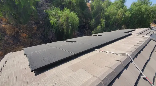 Black solar modules on a flat concrete tile roof, with conduit running over the ridge and trees on a slope behind, Mission Hills