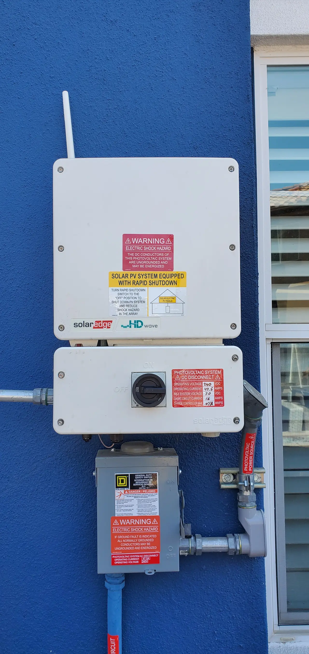 A SolarEdge HD-Wave inverter with its safety switch above an AC disconnect on a blue wall, Mission Hills
