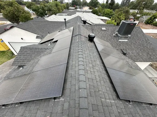 Solar modules on both sides of a shingle ridge with a turbine vent between them, neighboring roofs and trees beyond, Mission Hills