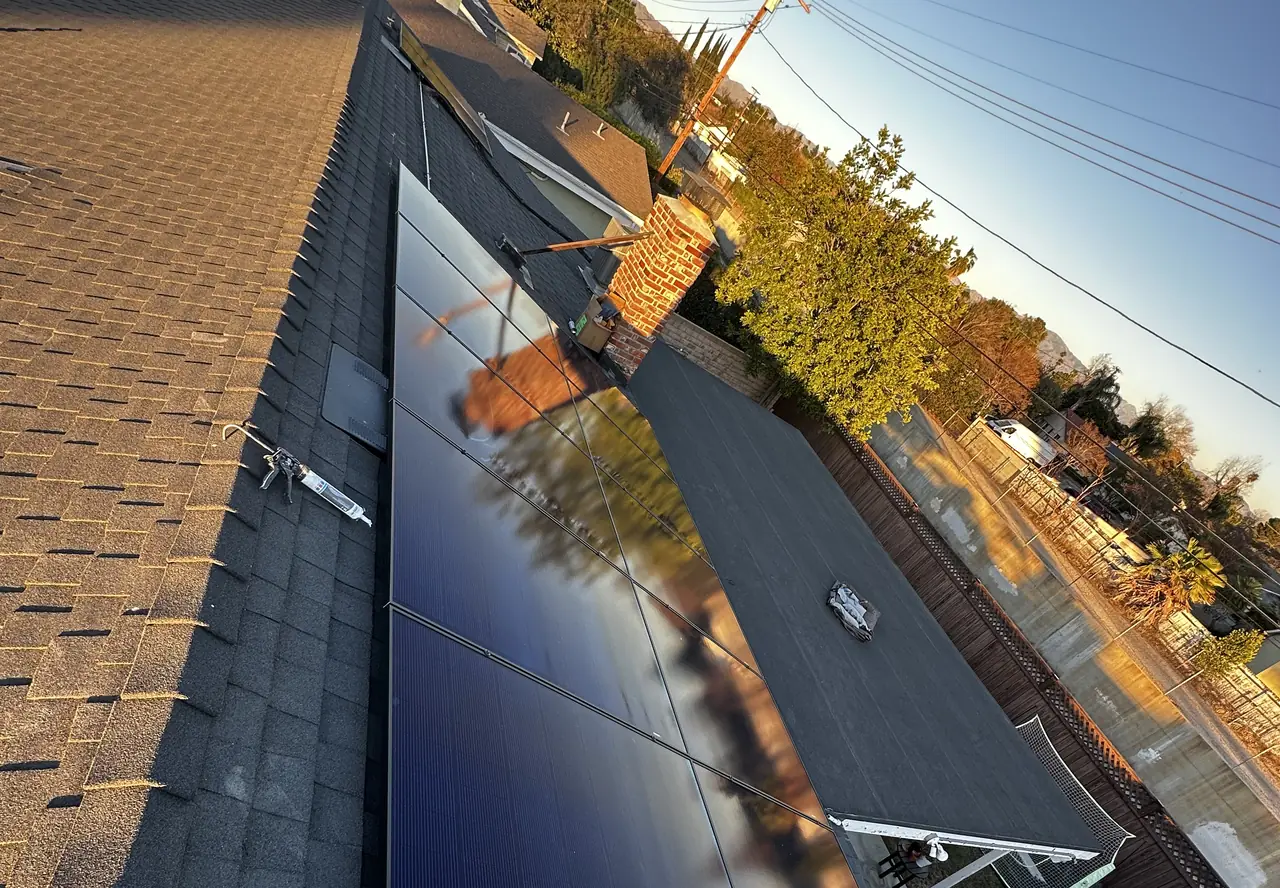 A solar module set at the edge of a shingle roof at sunset, with trees and overhead lines behind it, North Hills, Los Angeles