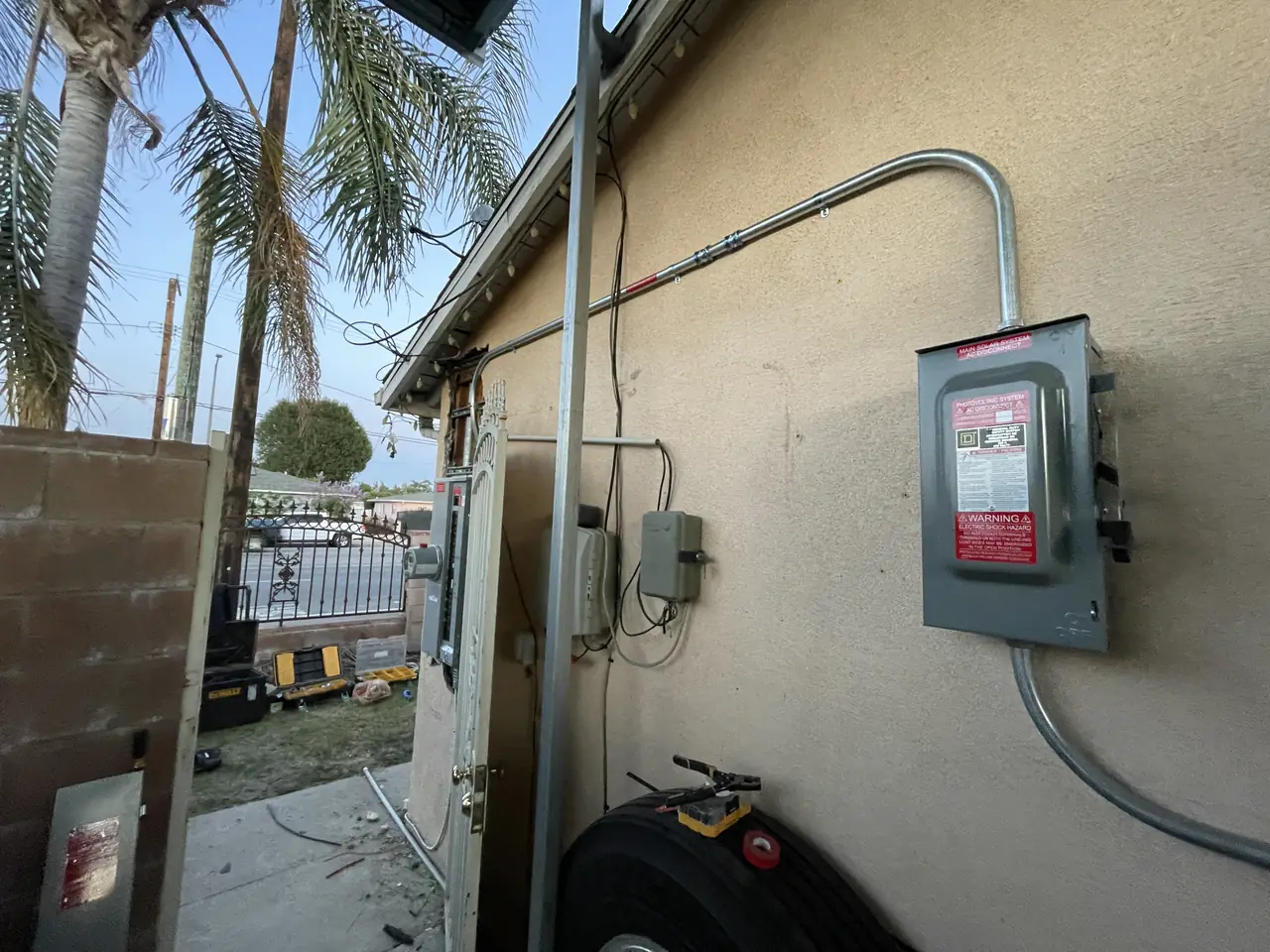 A stucco wall carrying a solar AC disconnect and conduit at dusk, with palms behind the house, North Hills, Los Angeles