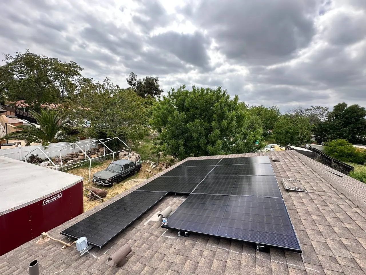 A solar array on a shingle roof under a cloudy sky with trees and a wide yard beyond, North Hills, Los Angeles