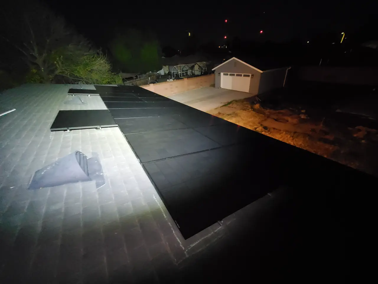 A solar array lit by a work light on a roof at night with a detached garage behind, North Hills, Los Angeles