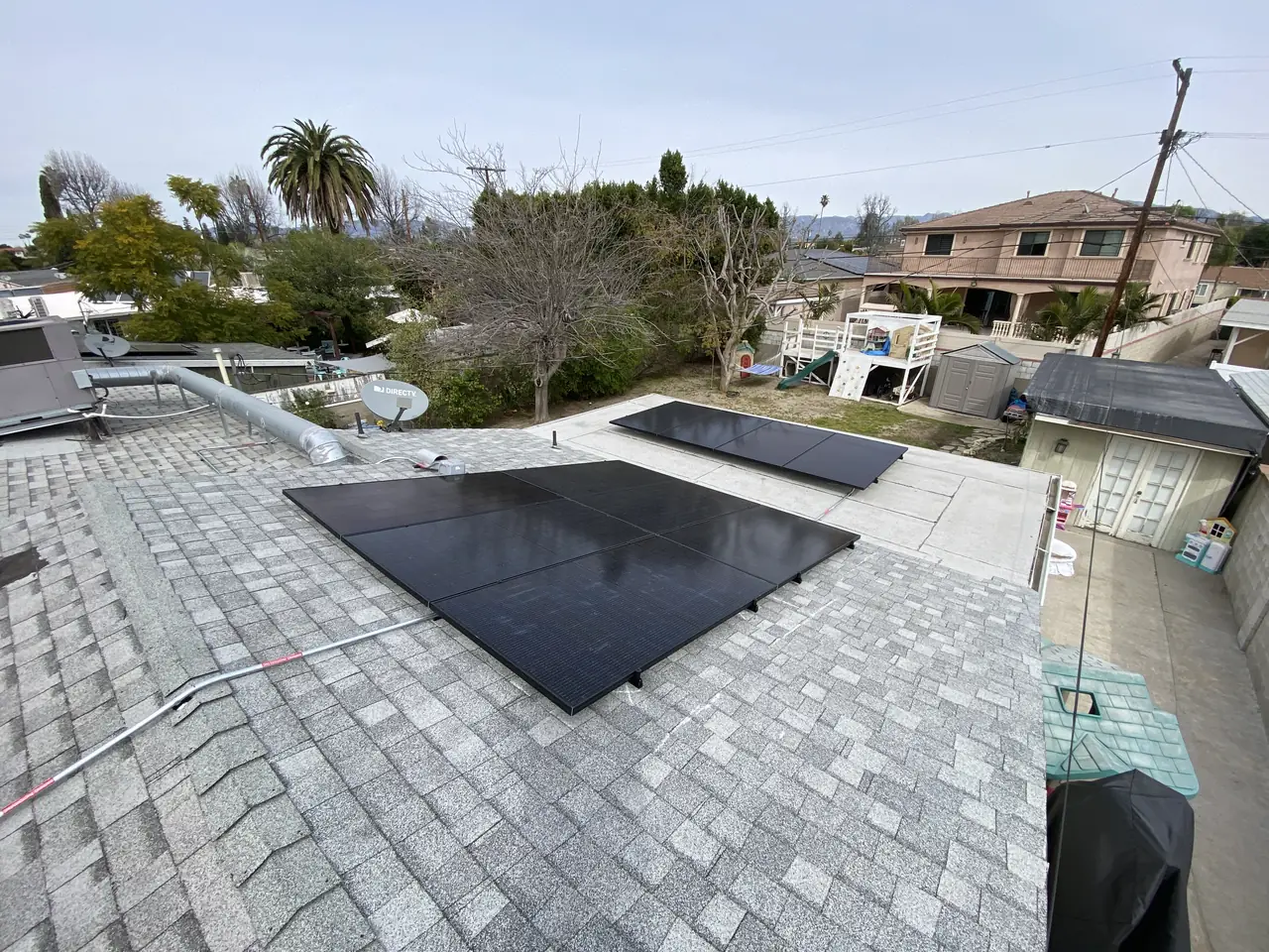 Two groups of solar modules across a shingle roof with neighbouring houses and a palm beyond, North Hollywood
