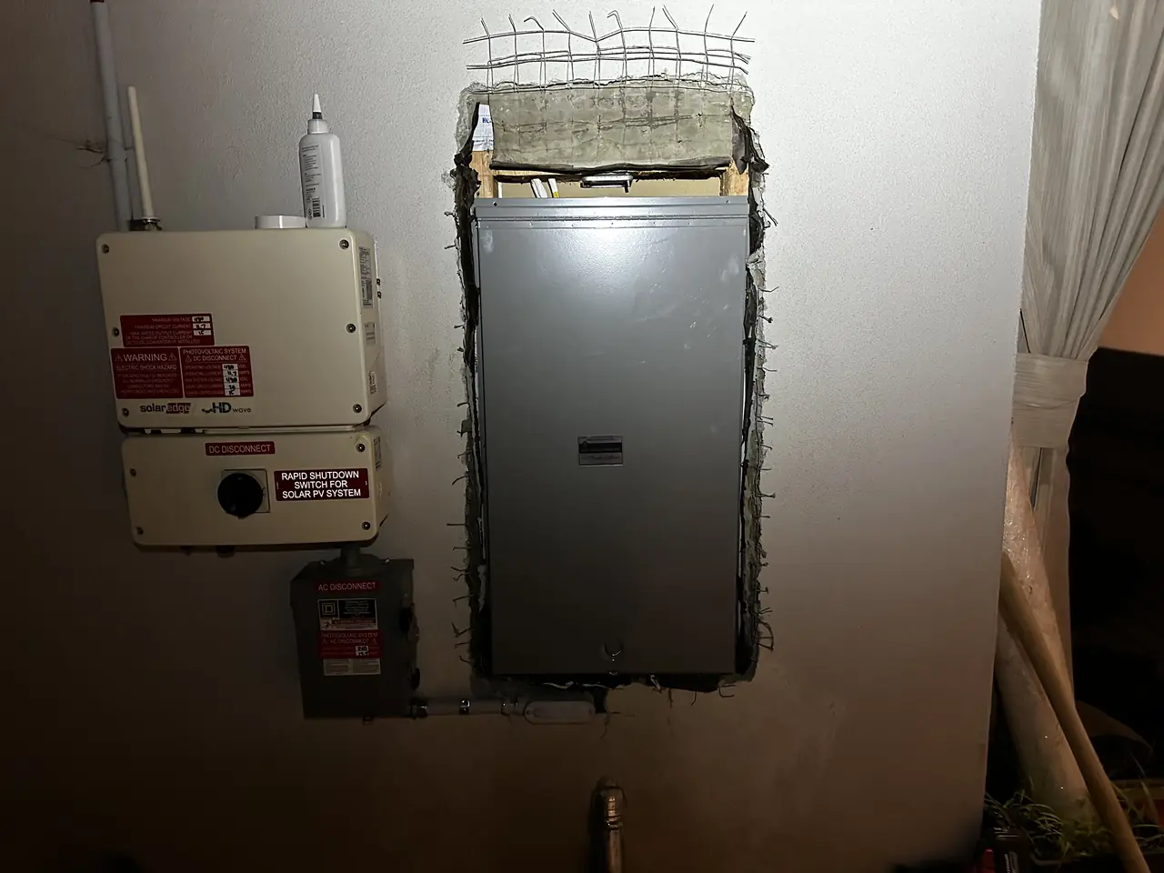 String inverter with a rapid-shutdown placard, an AC disconnect and the electrical panel on an exterior wall at night, North Hollywood