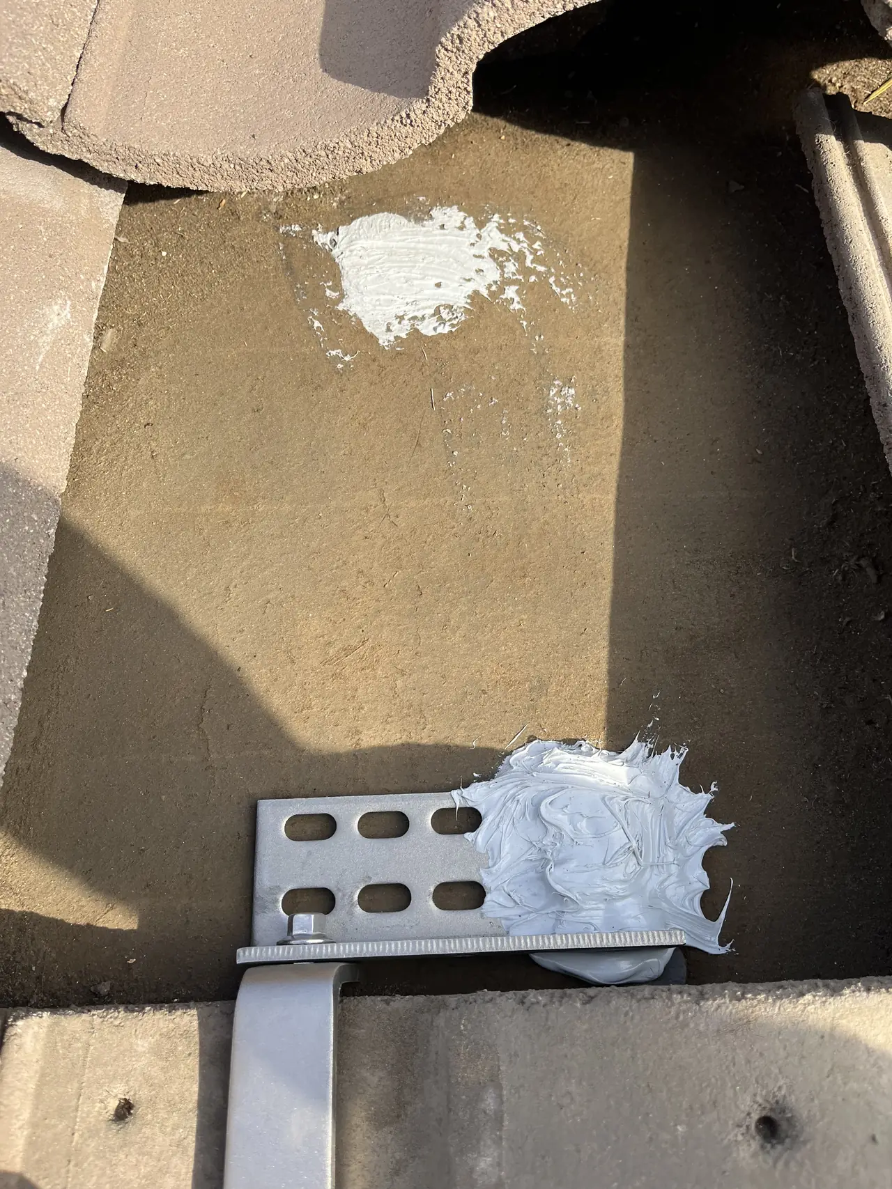 A slotted metal base plate lag-bolted through a concrete tile, with sealant over the fixings and the hook leg bent down from it, North Hollywood