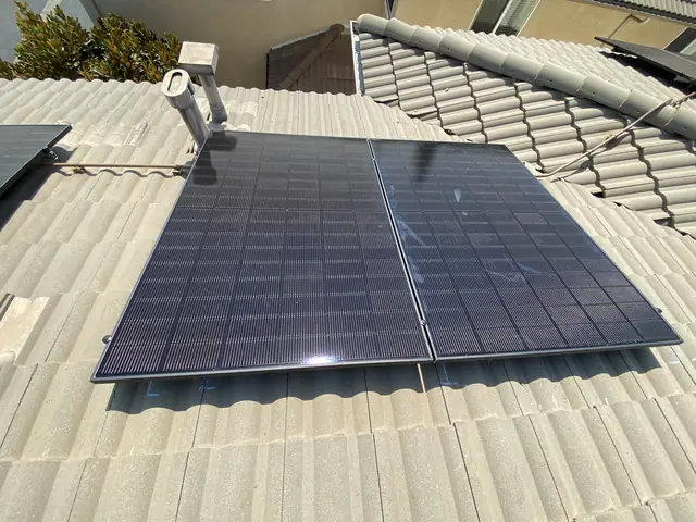 All-black solar modules laid across a pale gray concrete tile roof beside a stucco wall, Pacoima, Los Angeles