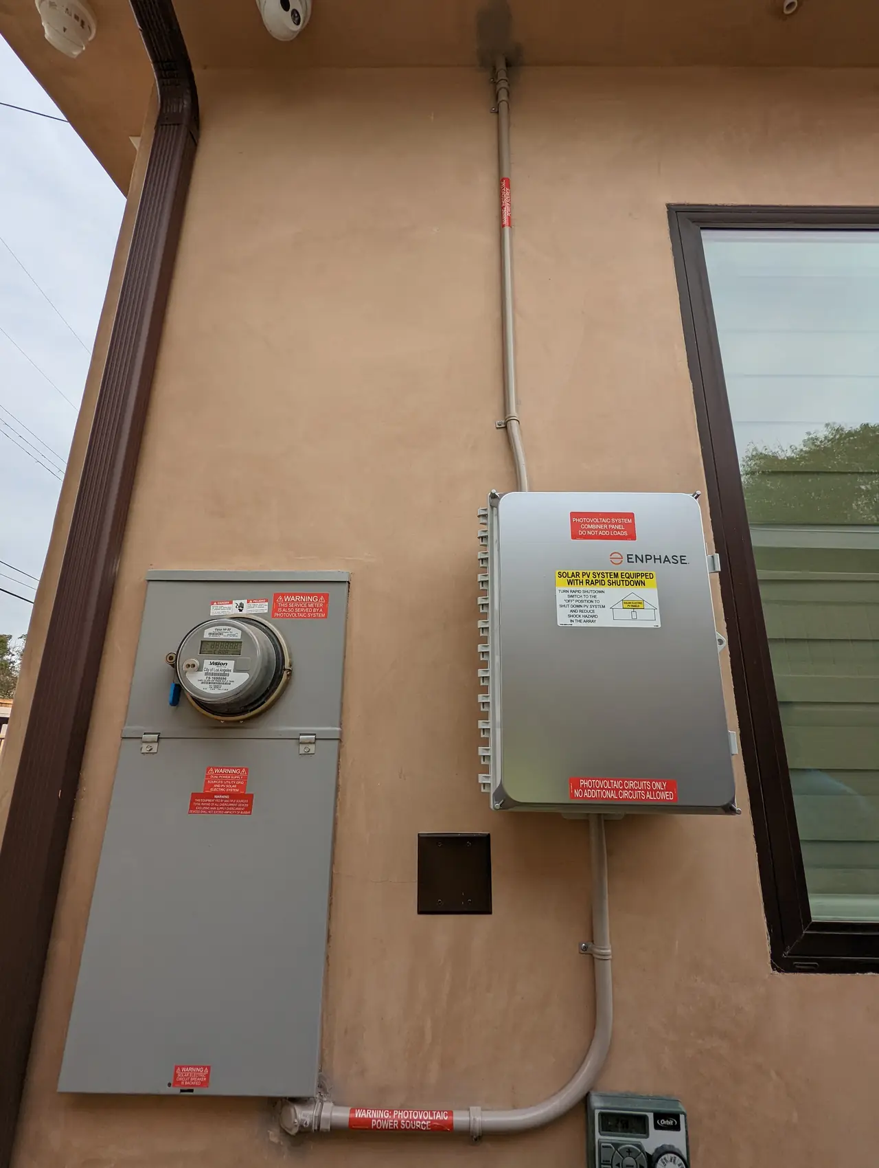 A meter enclosure and a closed Enphase combiner panel mounted side by side on a tan stucco wall, Panorama City