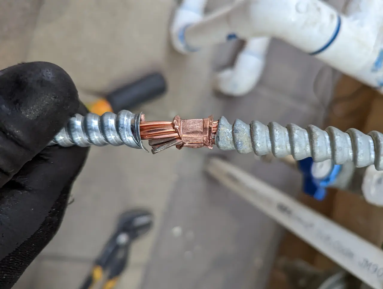 A gloved hand crimping a bare copper bonding conductor into a fitting on flexible metal conduit, Panorama City