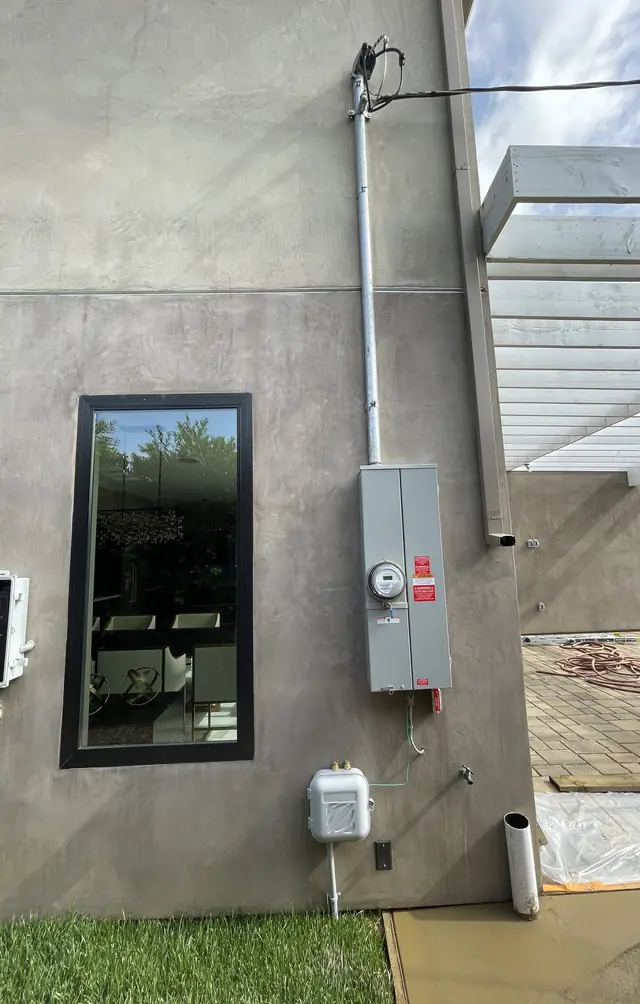 A meter and service enclosure on a gray stucco wall beside a window, with a white pergola beyond, Panorama City