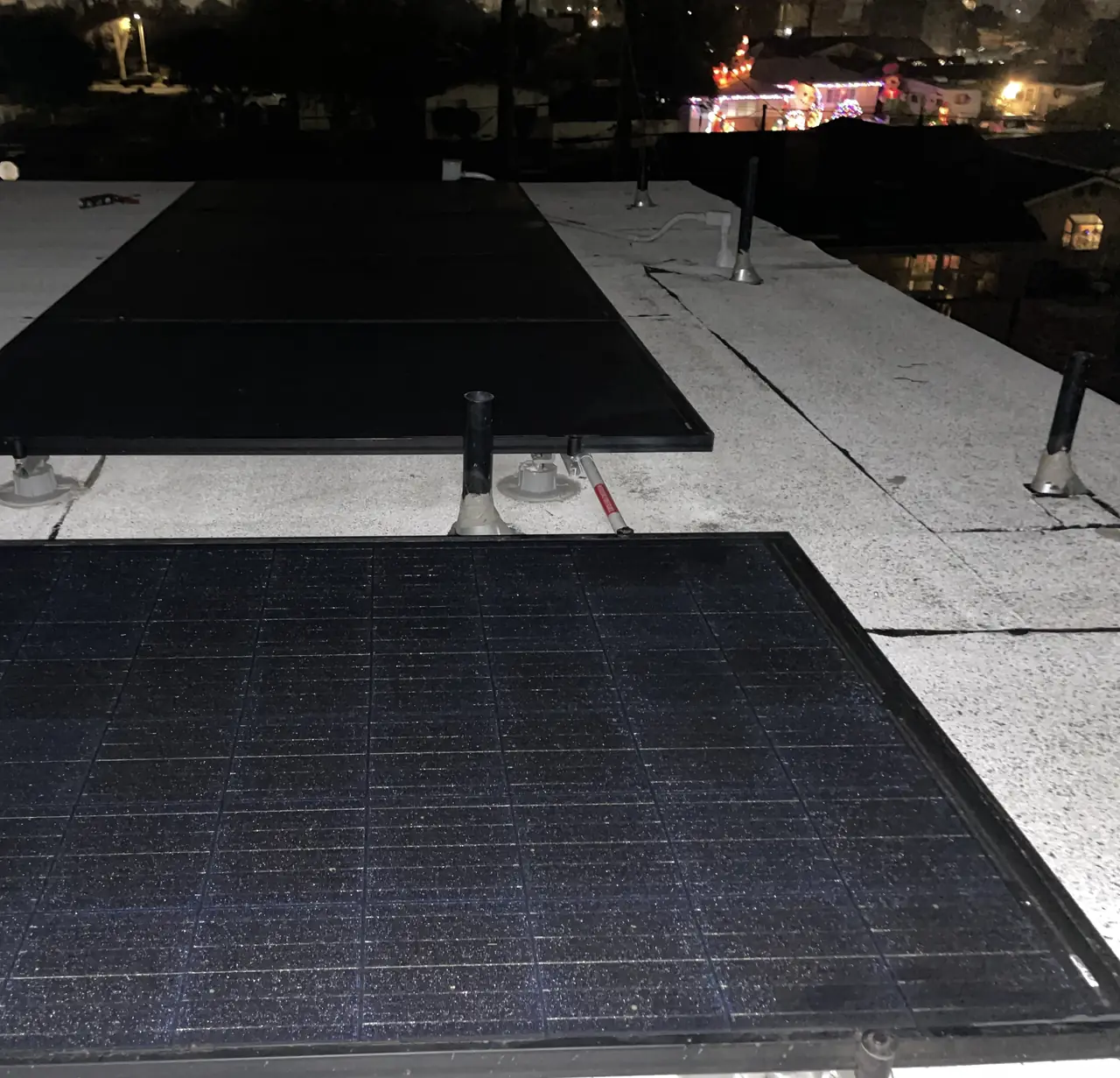 Two rows of black solar modules set close to horizontal on a white low-slope roof at night, with the neighborhood lit beyond, Panorama City