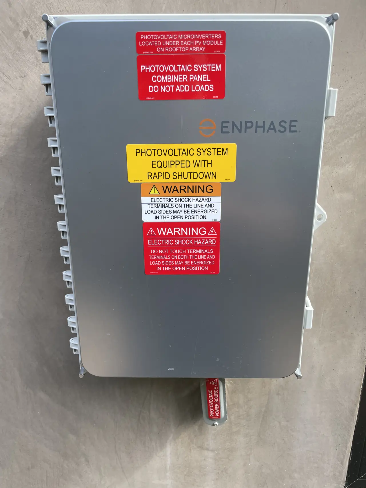 A closed Enphase combiner panel on a stucco wall carrying rapid shutdown and electric shock hazard placards, Panorama City