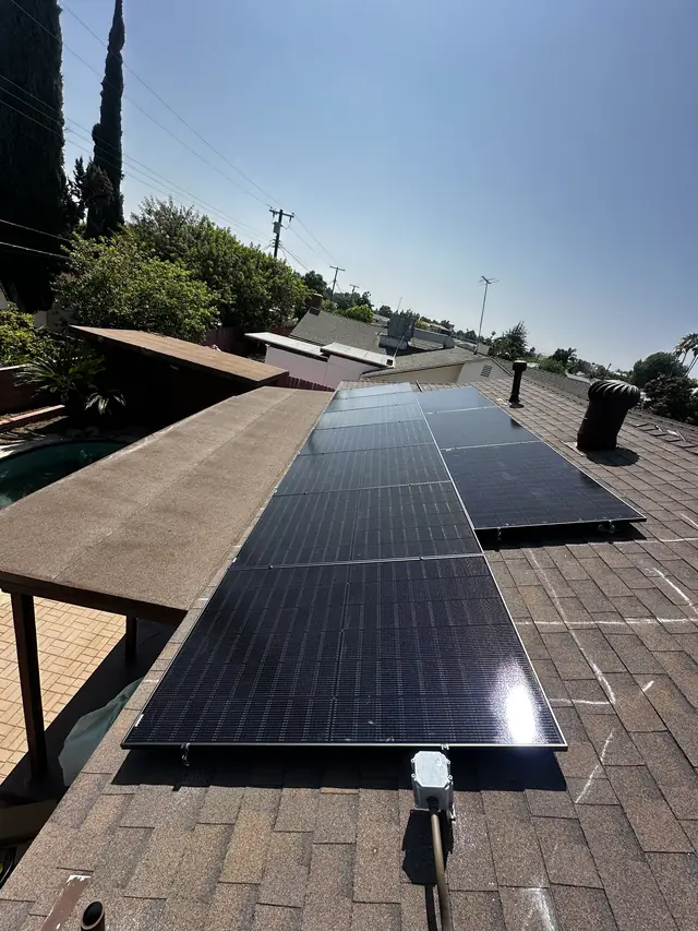 A long run of black solar modules along a composition shingle roof with the neighborhood and trees beyond, Panorama City