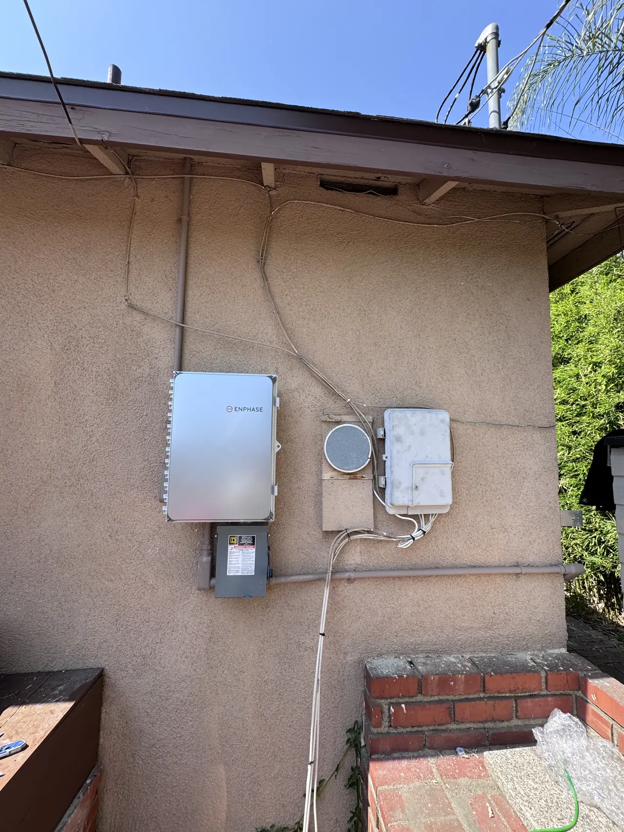A closed Enphase combiner and a fused disconnect mounted on a tan stucco wall beside older enclosures under the eaves, Panorama City