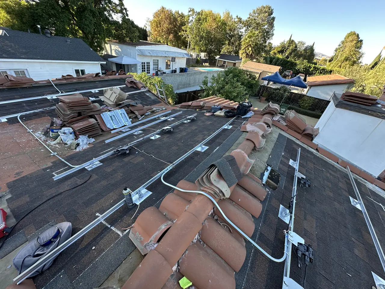 Roof with clay tile stripped back to a rectangle of asphalt shingle, mounting rails laid out, and the removed tile stacked around the edges, Reseda