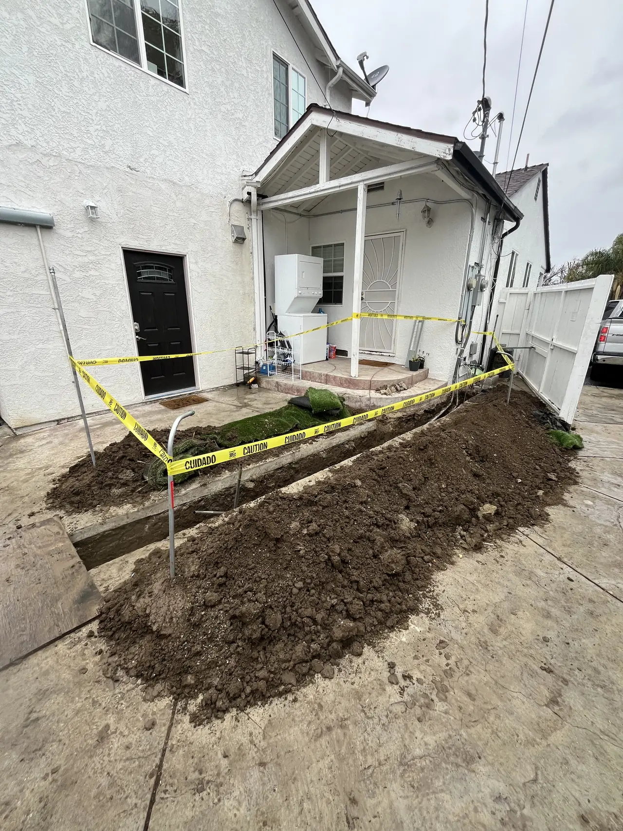 Open trench with excavated soil and yellow caution tape running along the side of a stucco house toward a covered porch, Reseda