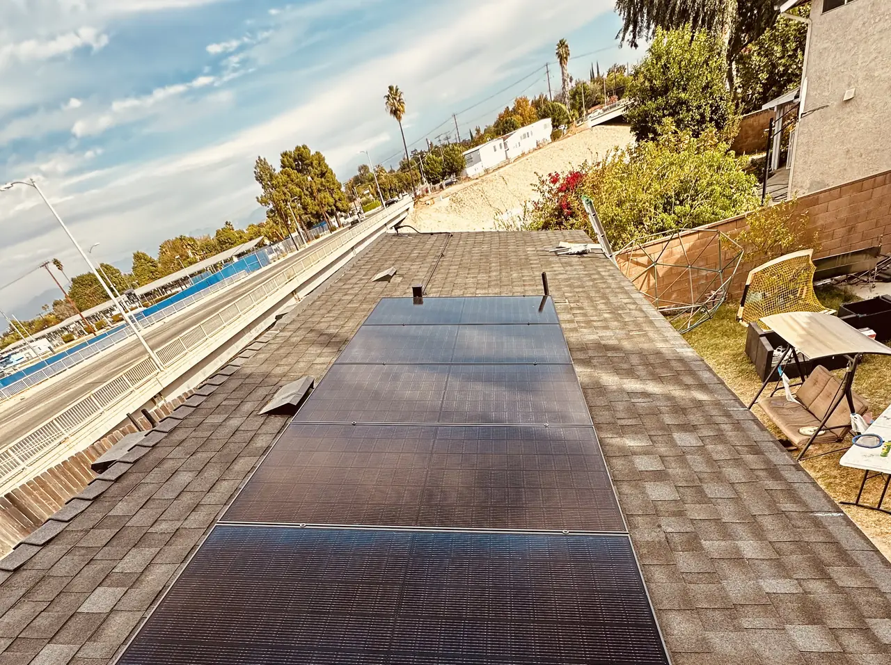 A single row of black solar modules on a brown composition shingle roof with palm trees and open sky beyond, Reseda