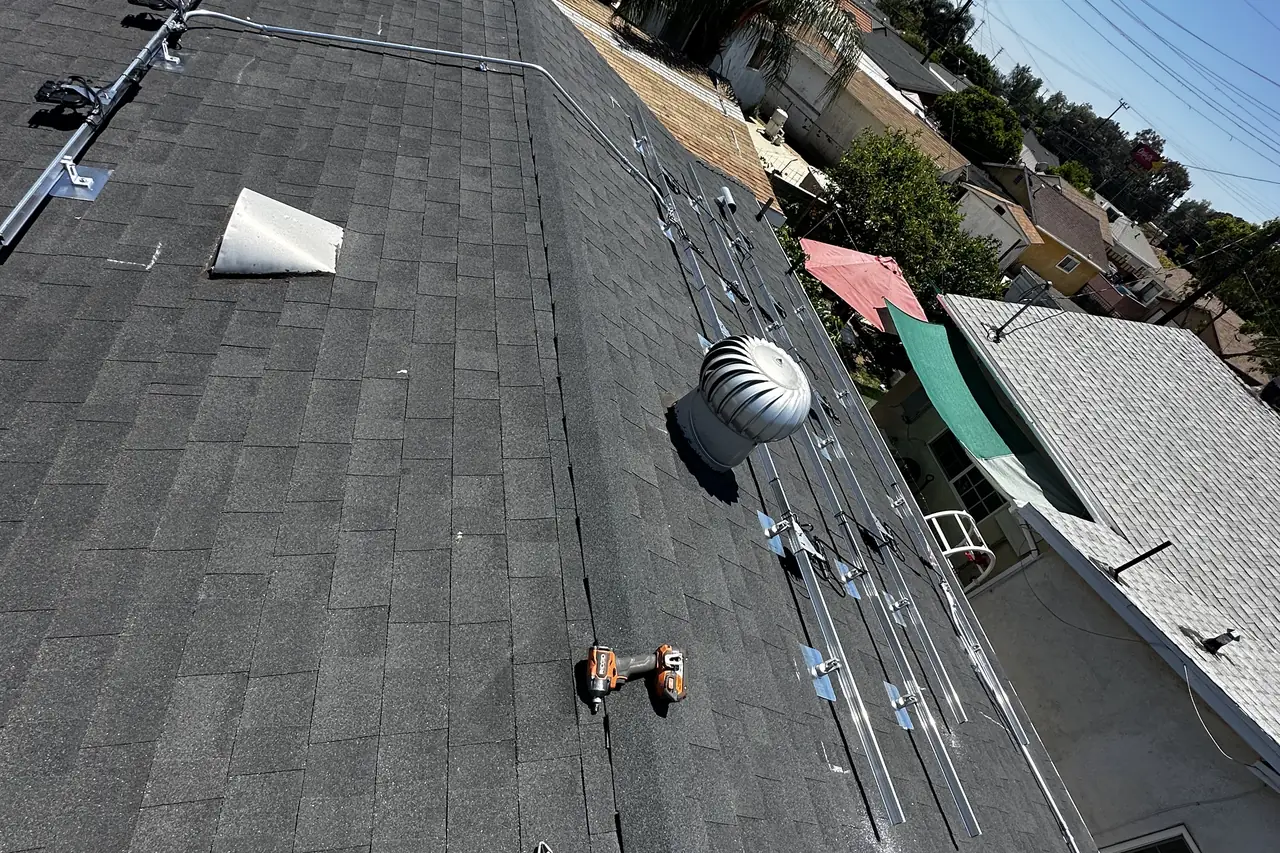 Aluminum solar rails laid across a dark shingle roof beside a turbine vent, with the conduit run along the top, San Fernando