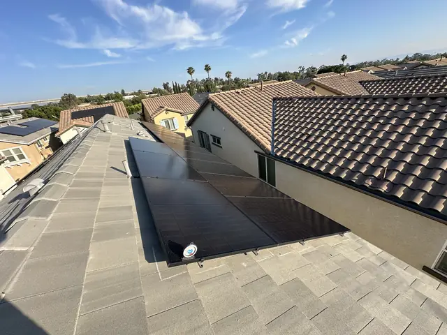 A row of black solar modules on a flat gray concrete tile roof, with neighboring two-story houses and clay tile roofs beyond, San Fernando