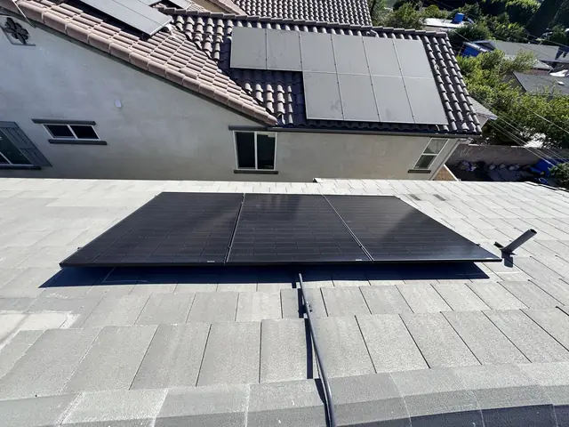 Black solar modules on a flat gray concrete tile roof, with a neighboring house and its solar array on clay tile behind, San Fernando
