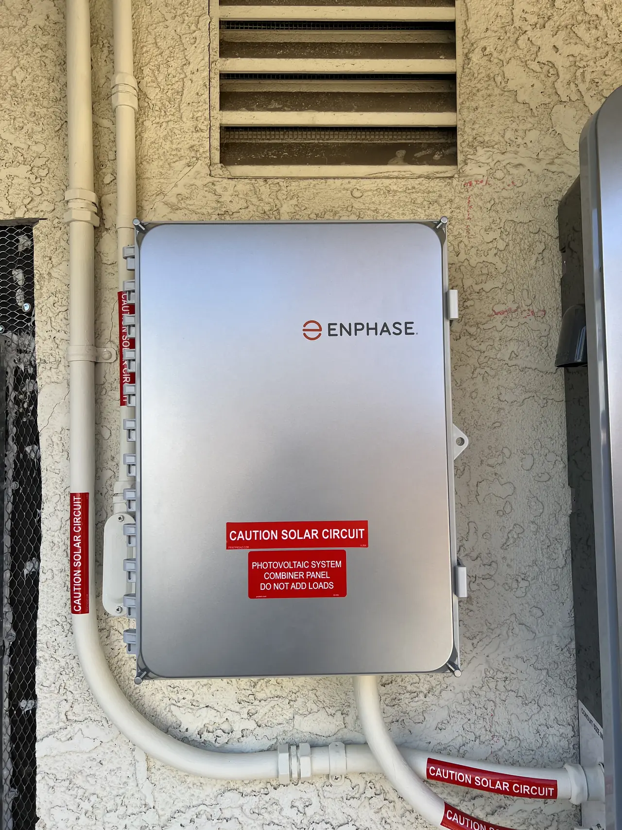 A closed combiner enclosure on a stucco wall below a vent, Santa Clarita