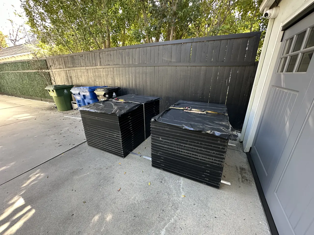 Pallets of solar modules wrapped in black film standing on a driveway beside a garage door, Studio City