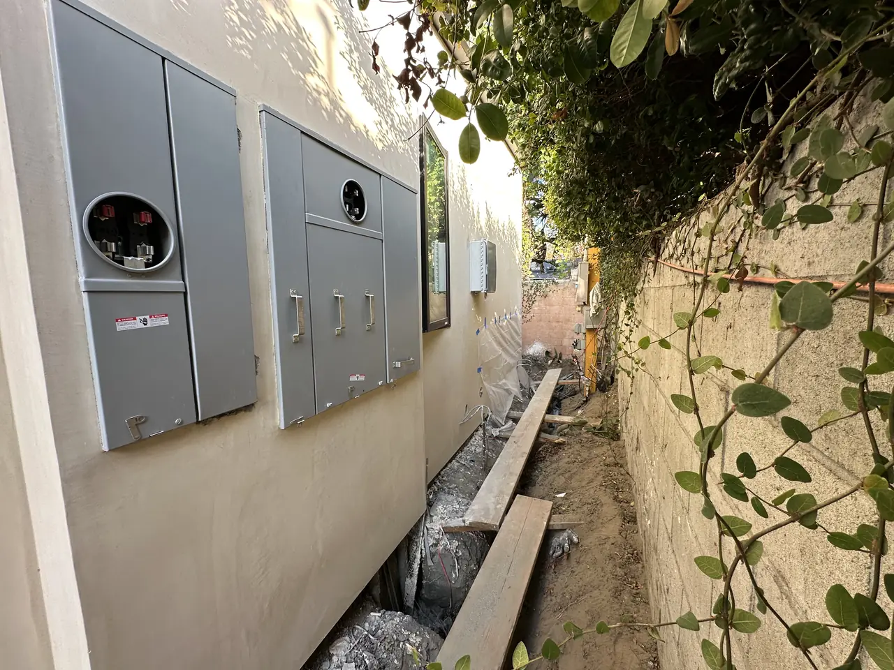 Meter socket and electrical enclosures on a finished stucco wall beside a planted retaining wall, Studio City